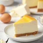"High-protein Greek yogurt cheesecake slice on a white plate with cream cheese, vanilla protein powder, and key ingredients around on a marble surface"