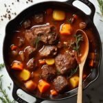 "Overhead shot of a rustic beef stew with chuck roast, turnips, parsnips, and herbs in a Dutch oven, garnished with sprigs of thyme and rosemary, on a white marble countertop with scattered herbs and crackled black pepper."