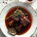 "Braised short ribs on white ceramic plate with rosemary, thyme, wine braising liquid, wooden spoon, and cracked black pepper"