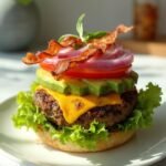 "Overhead shot of a bunless burger with beef patty, melted cheddar, avocado, tomato, and bacon nestled between lettuce and a grilled portobello mushroom cap on a clean marble countertop"