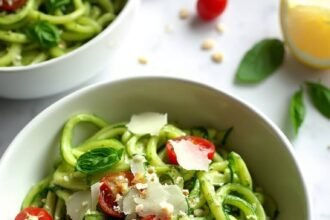 "Overhead shot of creamy pesto zucchini noodles garnished with cherry tomatoes, basil leaves, and Parmesan cheese in a white bowl on a marble surface"