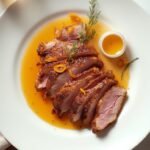 "Sliced crispy duck breast with orange butter sauce on white porcelain plate, garnished with orange zest and microgreens on a clean marble background"