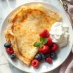 "Golden coconut flour crepes with fresh berries and whipped cream on a white porcelain plate, presented on a clean marble background"