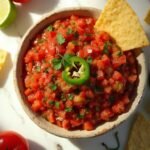 "Fresh homemade salsa with diced tomatoes, red onion, jalapenos, cilantro, and lime in a ceramic bowl on a white marble countertop, surrounded by tortilla chips, limes, and fresh ingredients."