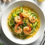 "Overhead shot of garlic butter shrimp scampi with zucchini noodles in a white ceramic bowl, garnished with parsley, lemon zest, and red pepper flakes on a marble background"