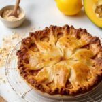 "Keto apple pie with a golden-brown almond flour crust on a cooling rack, with key ingredients like chayote squash, cinnamon, almond flour, and lemon in the background."