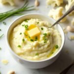 "Overhead shot of creamy mashed cauliflower garnished with chives and butter in a white ceramic bowl, surrounded by fresh ingredients on a marble countertop."