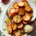 "Overhead view of roasted radishes with crispy edges, garlic butter, and rosemary on a white platter with Parmesan dusting, chopped chives, lemon zest, wooden spoon, whole radishes, and garlic cloves in the background."
