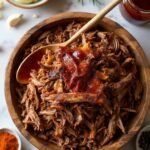 "Overhead shot of juicy slow-cooker pulled pork with caramelized edges in a rustic wooden bowl, with a jar of sugar-free BBQ sauce, garnishes and spices on a marble background"