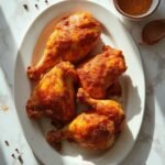 "Golden-brown crispy baked chicken thighs on a white platter with seasoning bowl on marble surface"