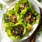 "Keto Asian beef lettuce wraps with cashews and water chestnuts on a white platter, garnished with green onion rings, cilantro leaves, and lime wedges, with a ramekin of chili garlic sauce on a marble countertop."