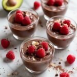 "Overhead view of dark chocolate avocado mousse in glass bowls garnished with raspberries and chocolate shavings on a white marble background"