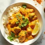"Overhead view of keto butter chicken over cauliflower rice in a white bowl on a marble background, with turmeric sauce, green cilantro, a lemon wedge, and scattered spices"