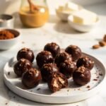 "Keto chocolate almond fat bombs on a white plate with almond butter, cocoa powder, and coconut oil in the background"
