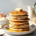 "Stack of golden-brown, fluffy coconut flour keto pancakes with melted butter on a white plate, surrounded by ingredients like coconut flour, eggshells, heavy cream, and vanilla beans, shot in warm natural light."