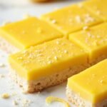 "Sugar-free keto lemon bars with almond crust arranged on a clean white marble background, dusted with powdered erythritol, and accompanied by fresh lemon zest and halved lemons."