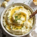 "Overhead shot of creamy keto mashed cauliflower in a white bowl, garnished with chives and Parmesan, with a wooden spoon, cauliflower florets, and garlic cloves arranged around it."