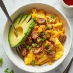"Keto sausage and egg breakfast bowl with cheddar cheese, garnished with scallions and avocado on a clean marble background"