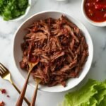 "Slow cooker keto beef barbacoa in a white bowl surrounded by toppings like lime wedges, onions, chipotle peppers, and lettuce cups, on a white marble surface."