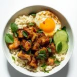 "Thai basil chicken over cauliflower rice with lime, cucumber, and a fried egg in a white ceramic bowl"