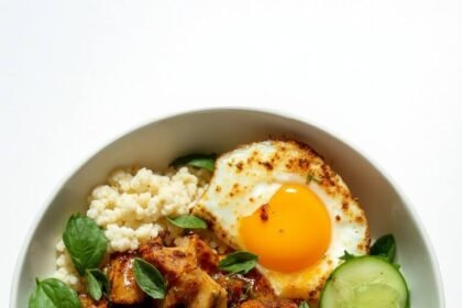 "Thai basil chicken over cauliflower rice with lime, cucumber, and a fried egg in a white ceramic bowl"